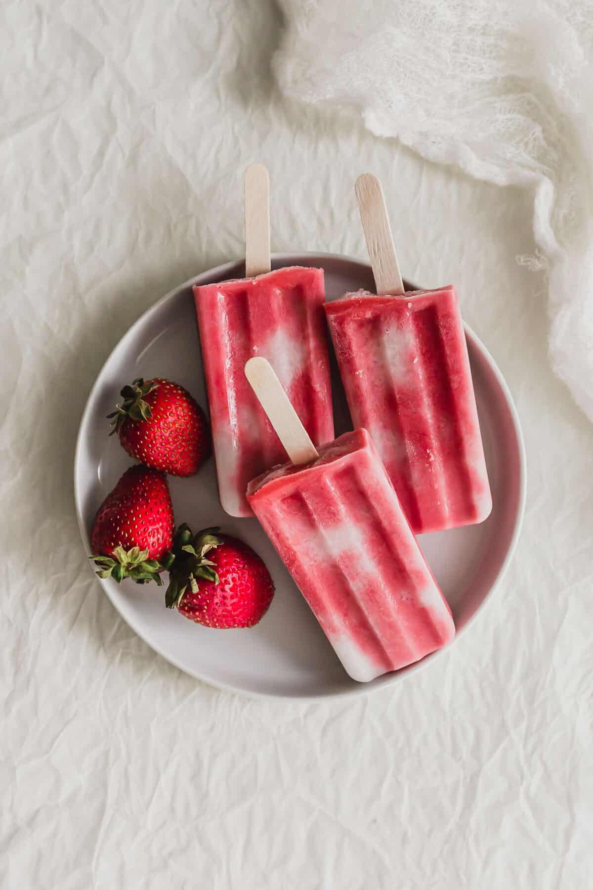 3 Strawberry sago popsicles piled on a white plate, with strawberries on the side.